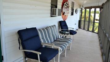 Une véranda couverte avec un plancher en bois peint en blanc, un canapé bleu avec des coussins, un coussin à rayures et une petite table ronde.