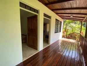 A wooden deck with a covered area, a door leading to an interior room, and a view of greenery outside.