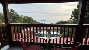 A balcony with a glass table and two wicker chairs, overlooking a coastal view with buildings and trees. - Stunning one bed/bath unit. Breathtaking ocean/jungle views with infinity pool! (Quepos)