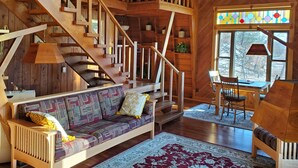 A wooden interior with a staircase, a sofa, a dining table, and a window with a view of trees.
