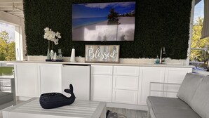 A modern outdoor seating area with a white sofa, a table with a decorative item, a flat-screen TV mounted on a green wall, and a sign that reads "Blessed."