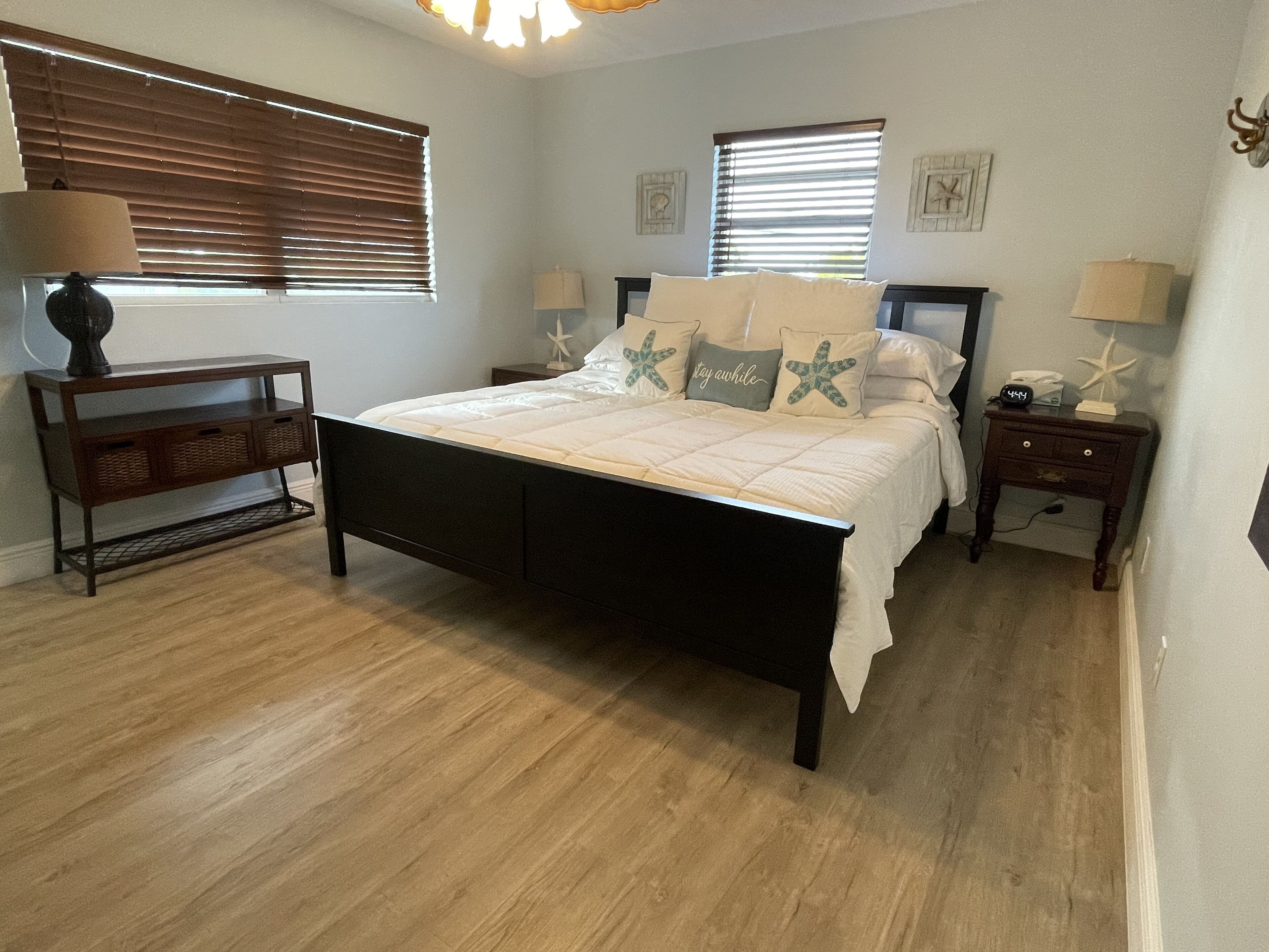 A bedroom with a bed, two bedside tables, a nightstand, a lamp, and a window with blinds.