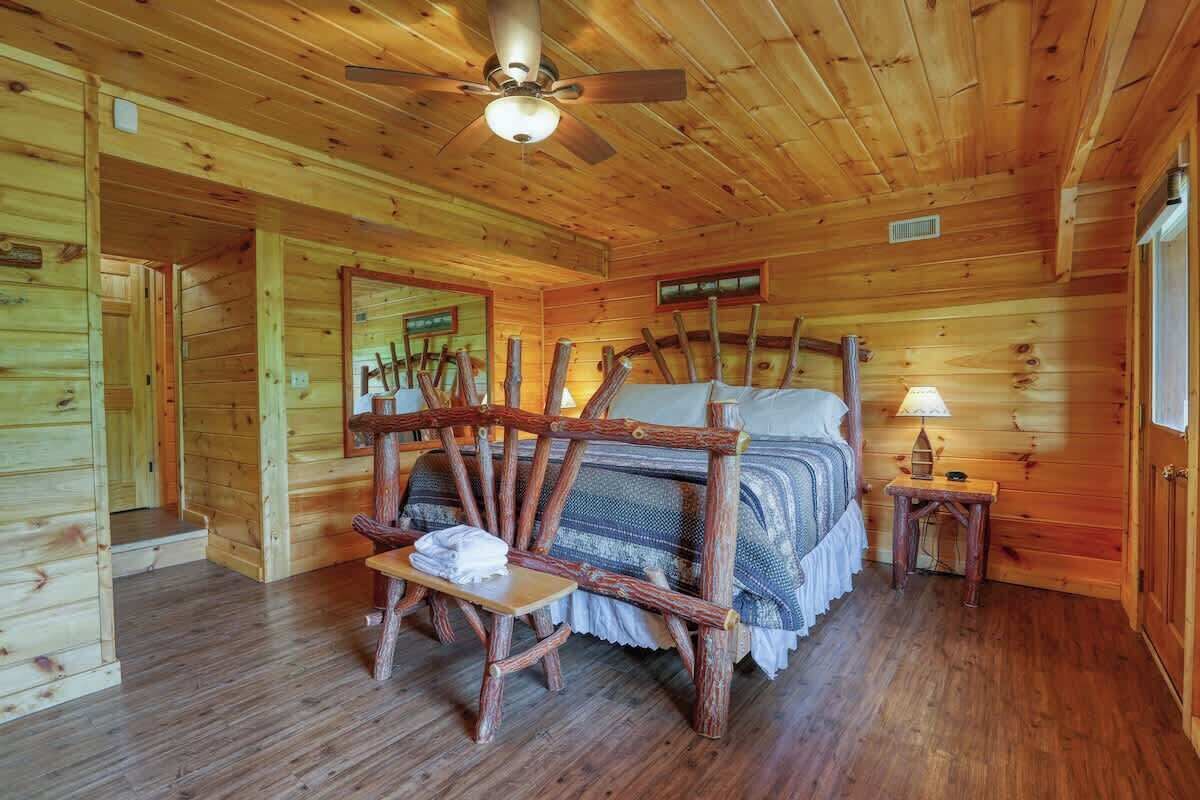 Quarto de cabana de madeira com uma cama, uma cadeira de madeira, uma mesa pequena e um ventilador de teto.
