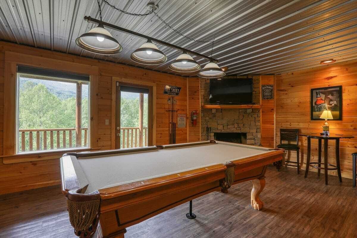 Una mesa de billar en una sala de madera con ventanas amplias, una chimenea y un televisor montado.