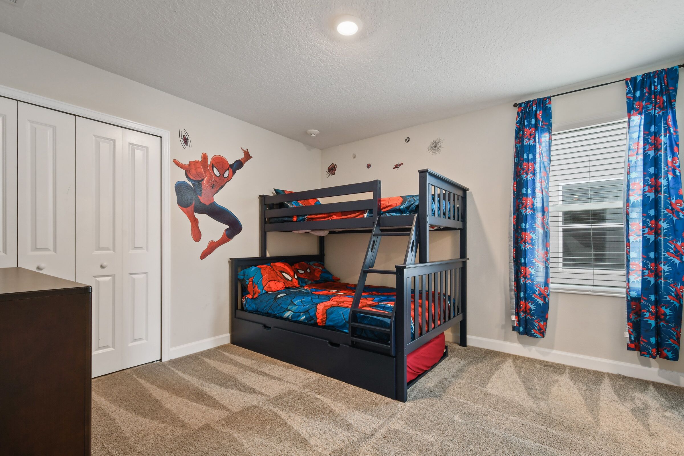 Un lit superposé avec une couette aux motifs de Spider-Man, une armoire blanche et une fenêtre ornée de rideaux à motifs floraux bleus.