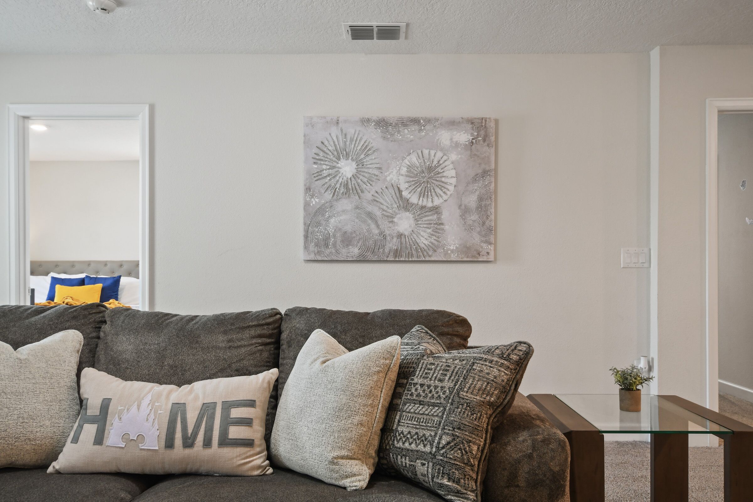 Un salon moderne avec un canapé gris, une table basse en verre et une œuvre d’art abstraite accrochée au mur.