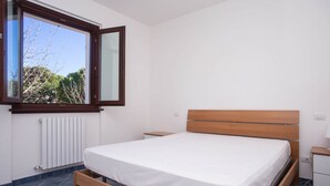 A bedroom with a wooden bed, a window with a view of trees, a white radiator, and a small bedside table. - M290 - Marcelli, new three-room apartment with terrace (Marcelli)