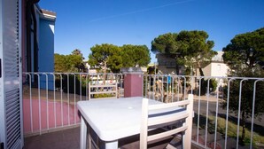 A balcony with a white table and chair, metal railing, and a view of trees and buildings. - M290 - Marcelli, new three-room apartment with terrace (Marcelli)