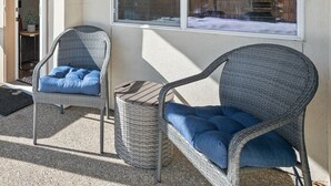 Un espacio de estar al aire libre techado con sillas de mimbre y una mesita, una ventana con persianas, y una puerta que da al interior.