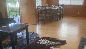 Een eetruimte met een houten tafel en stoelen, een leren vloerkleed en uitzicht op de omgeving door een glazen deur.