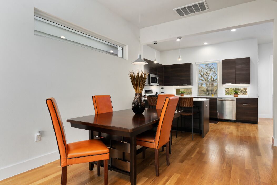 A modern kitchen with dark cabinets, a central island, and a dining area with a wooden table and orange chairs.