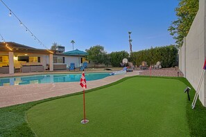 Un patio trasero con piscina, una casa pequeña y un área verde con una bandera.