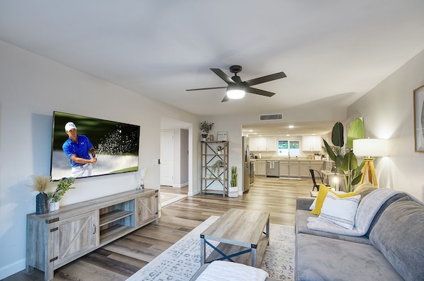 Un soggiorno moderno con una TV a schermo piatto, una ventola a soffitto e una cucina sullo sfondo.