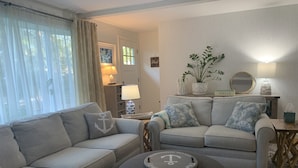 Una sala de estar con dos sofás, una mesa de centro y una mesita auxiliar con una lámpara. Se aprecia una ventana con cortinas, una puerta y una estantería de pared con objetos decorativos.