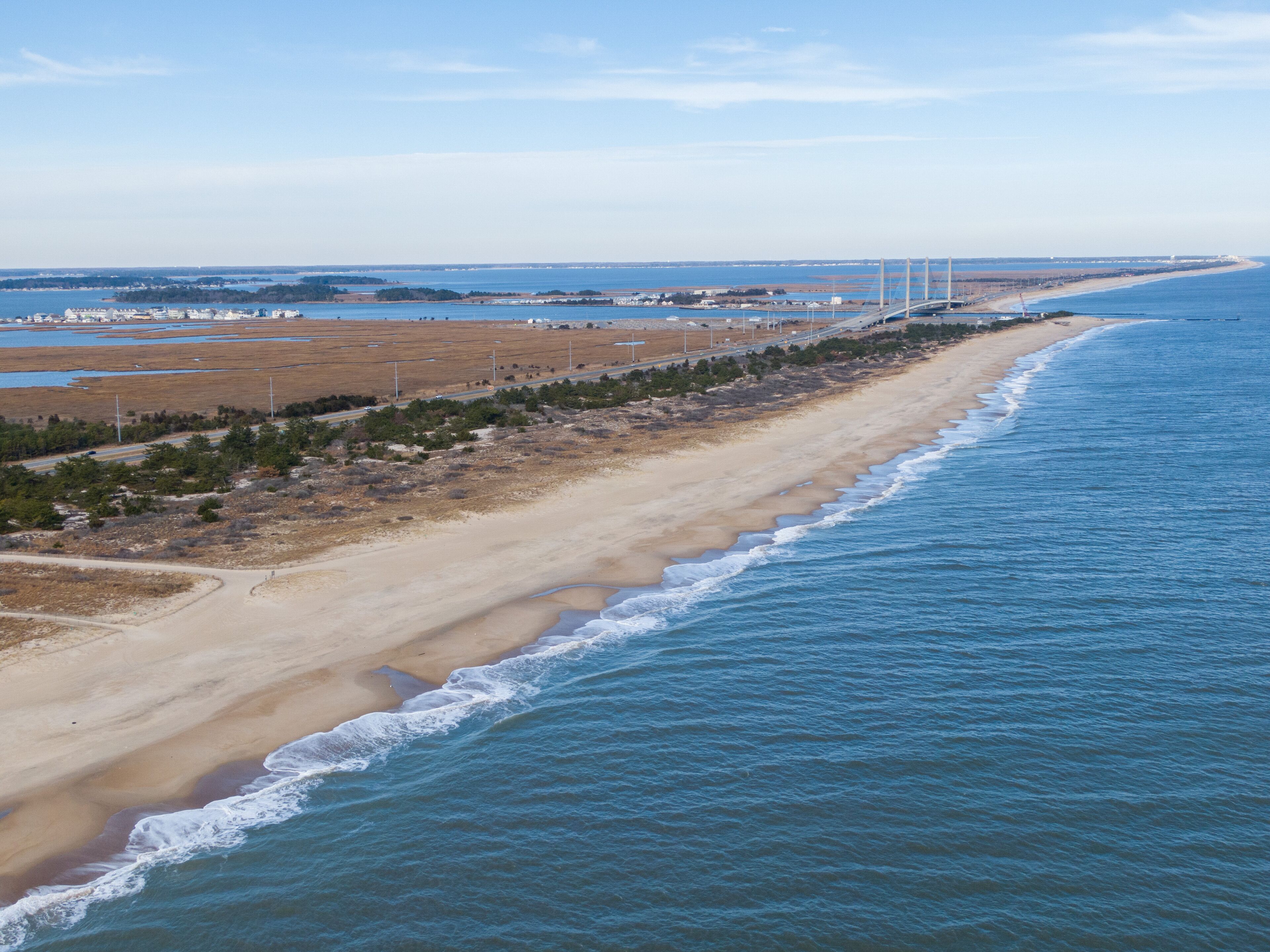 Eine Vogelperspektive auf eine Küstenregion mit einem Sandstrand, einer parallel zum Ufer verlaufenden Straße und einer Brücke in der Ferne.