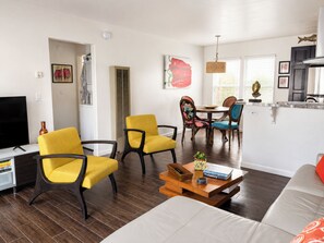 A modern living room with a flat-screen TV, yellow chairs, a white sofa, and a dining area with a table and chairs. - LA JOLLA SHORES PAD (LA JOLLA)