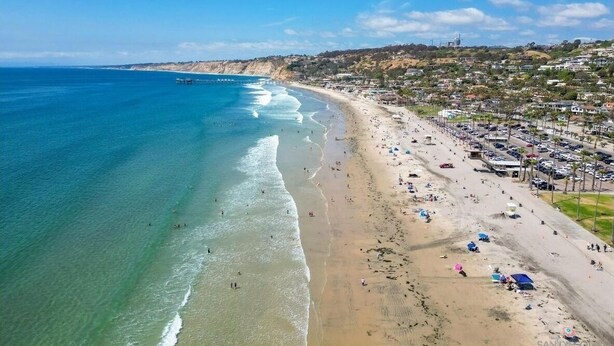 Una playa con aguas azules cristalinas, un muelle y una zona residencial con casas y coches aparcados.