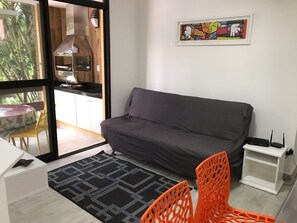 A modern living room with a grey sofa, a patterned rug, and an orange chair. A kitchen is visible through a glass door.