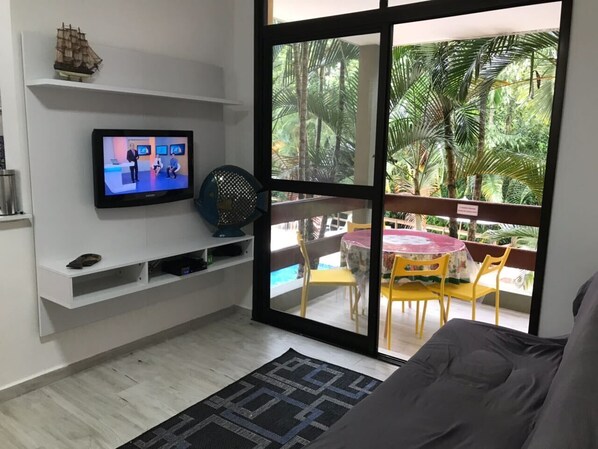 A living room with a television, a sofa, a small table, and a view of a pool and palm trees through a glass door.