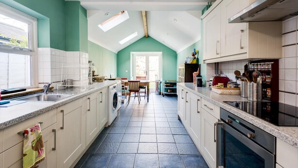 A kitchen with a central island, a dining area with a table and chairs, and a view of the outdoors through a door. - Lovely Terraced 2-bed House Near Hinksey Park (Oxford)