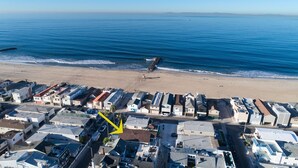 Vue aérienne d’une zone résidentielle côtière, avec des maisons le long de la plage, une digue rocheuse et une vue dégagée sur l’océan.