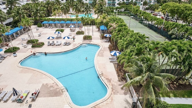 A large pool with lounge chairs and umbrellas, surrounded by palm trees and a tennis court.