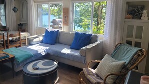A cozy living room with a sofa, a drum, a wicker chair, and a view of a lake through large windows.