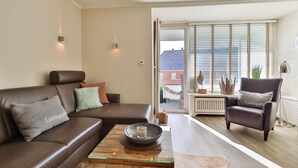 A modern living room with a brown leather sofa, a glass coffee table, and a leather armchair. There are two wall-mounted lights and a view of the outside through a sliding glass door.