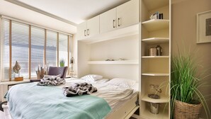 A bedroom with a bed, a large window with blinds, a bedside table, and a built-in shelving unit.