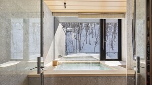 A modern spa room with a large bathtub, marble walls, and a view of a snowy forest through a large window.