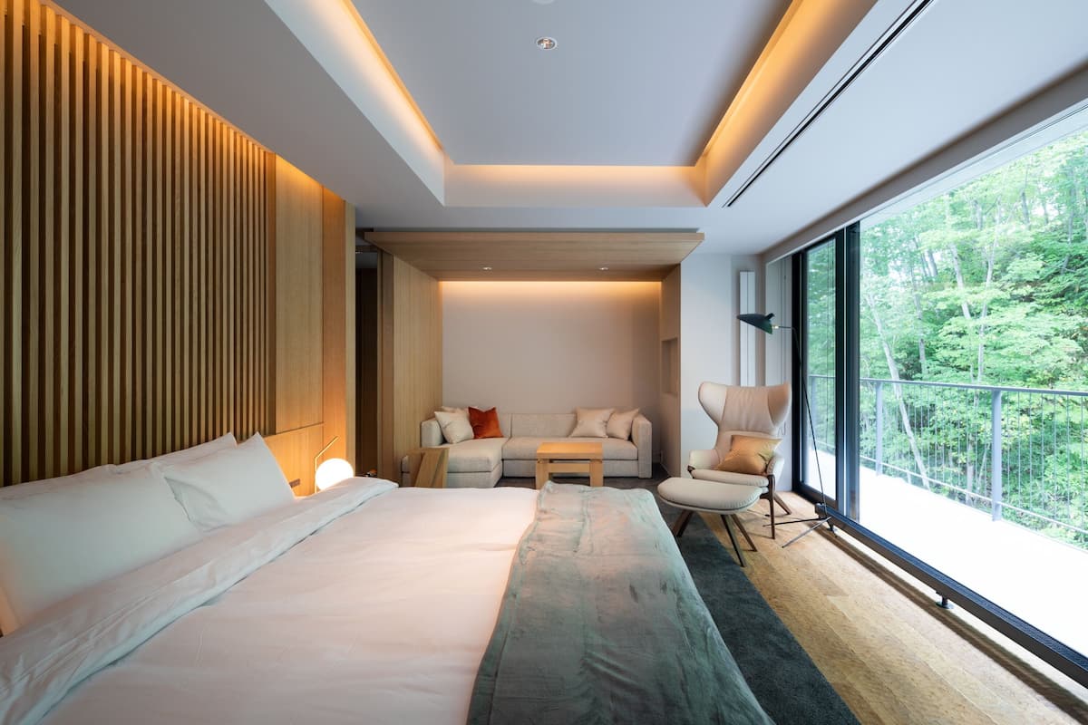 A modern hotel room with a large bed, a sofa, a chair, and a balcony with a view of the forest.