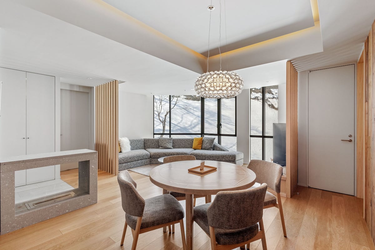 A modern living room with a round wooden table, grey upholstered sofa, and a large window with a view of a snowy landscape.