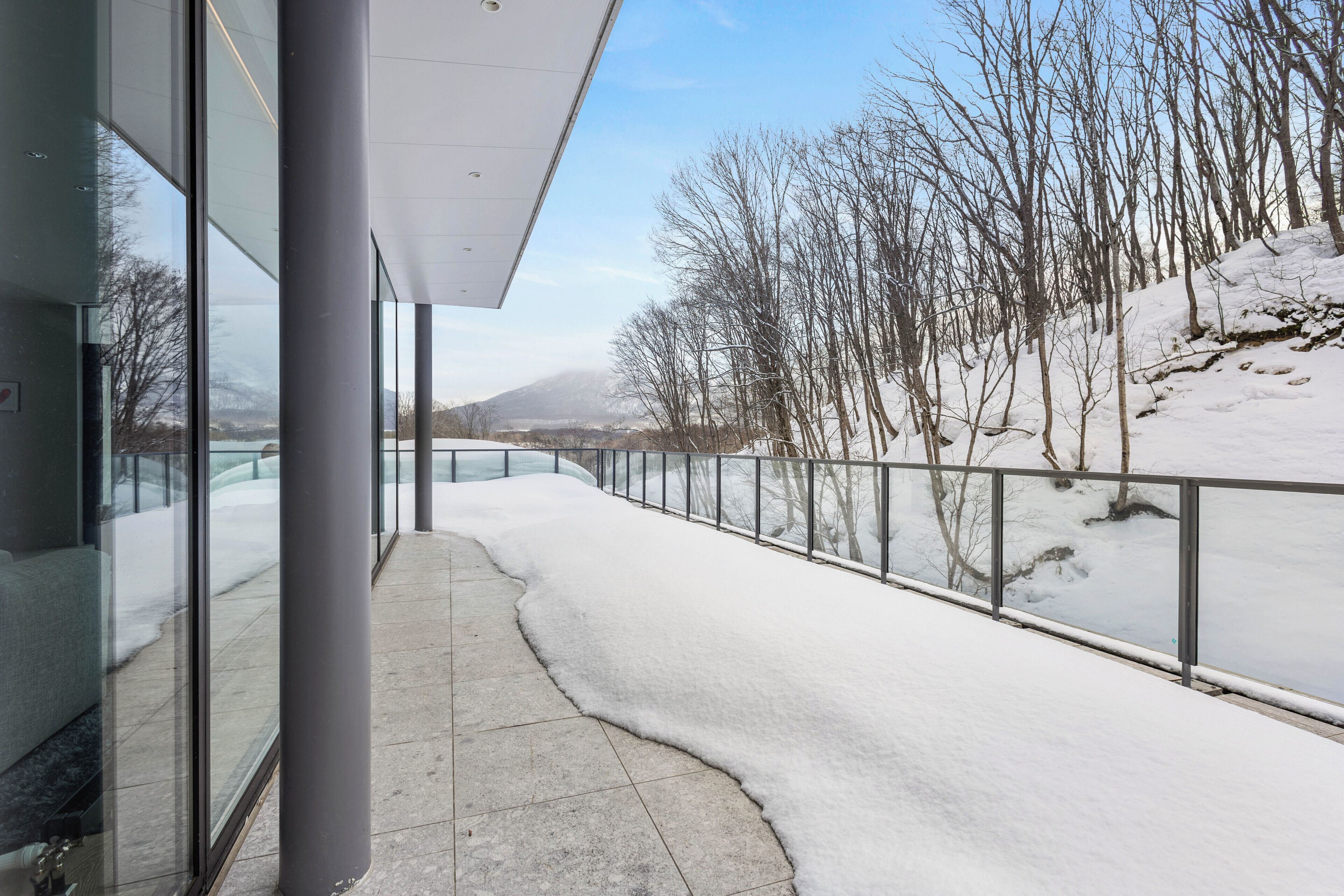 A snow-covered pathway leading to a building with large windows and a view of a snowy landscape with trees and a distant hill.