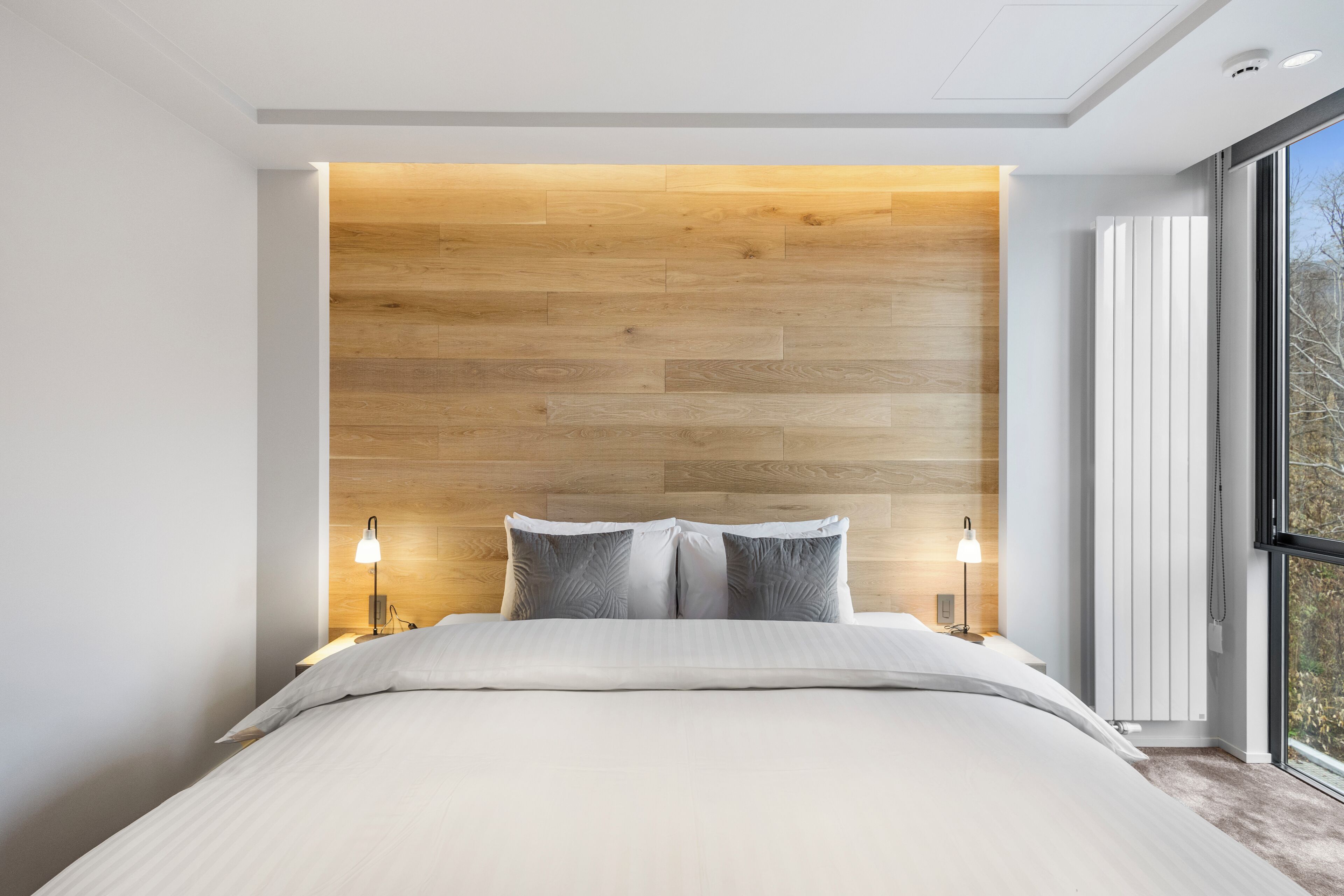 A modern bedroom with a wooden headboard, a large window, and a neatly made bed.