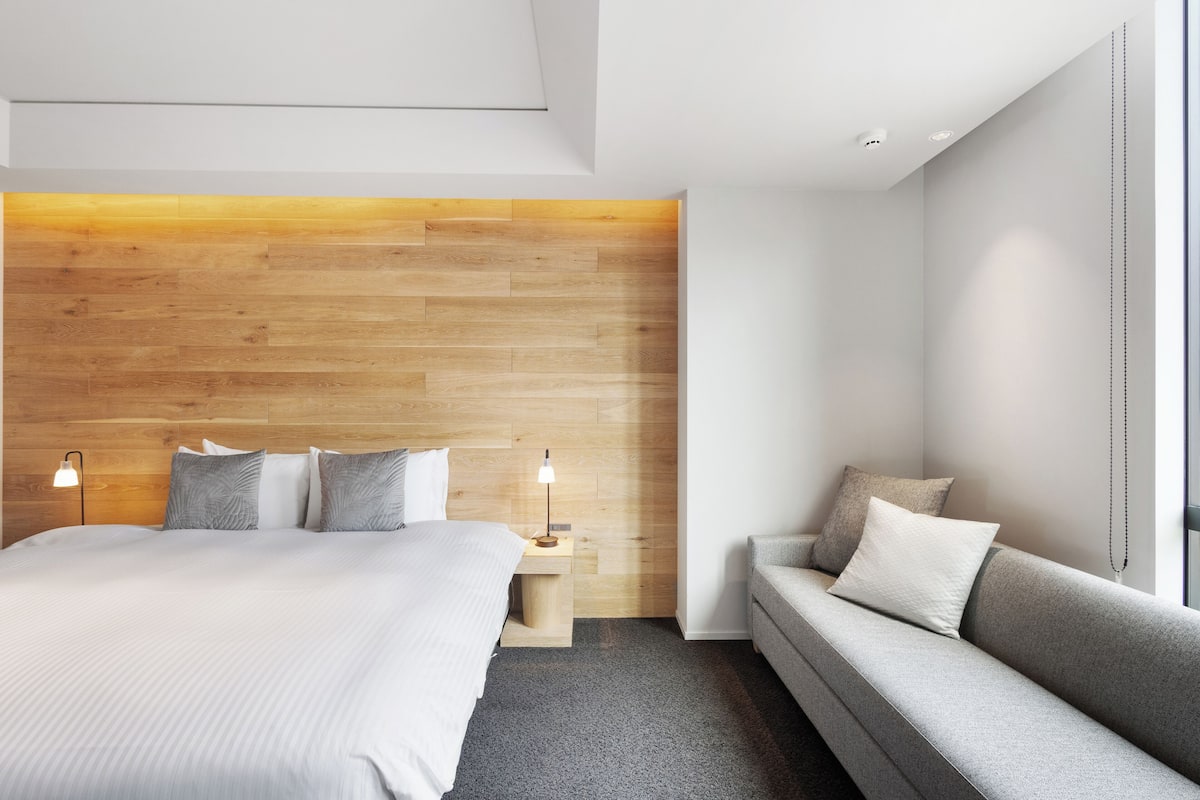 A modern hotel room with a wooden headboard, a large bed, a grey sofa, and a small bedside table with a lamp.