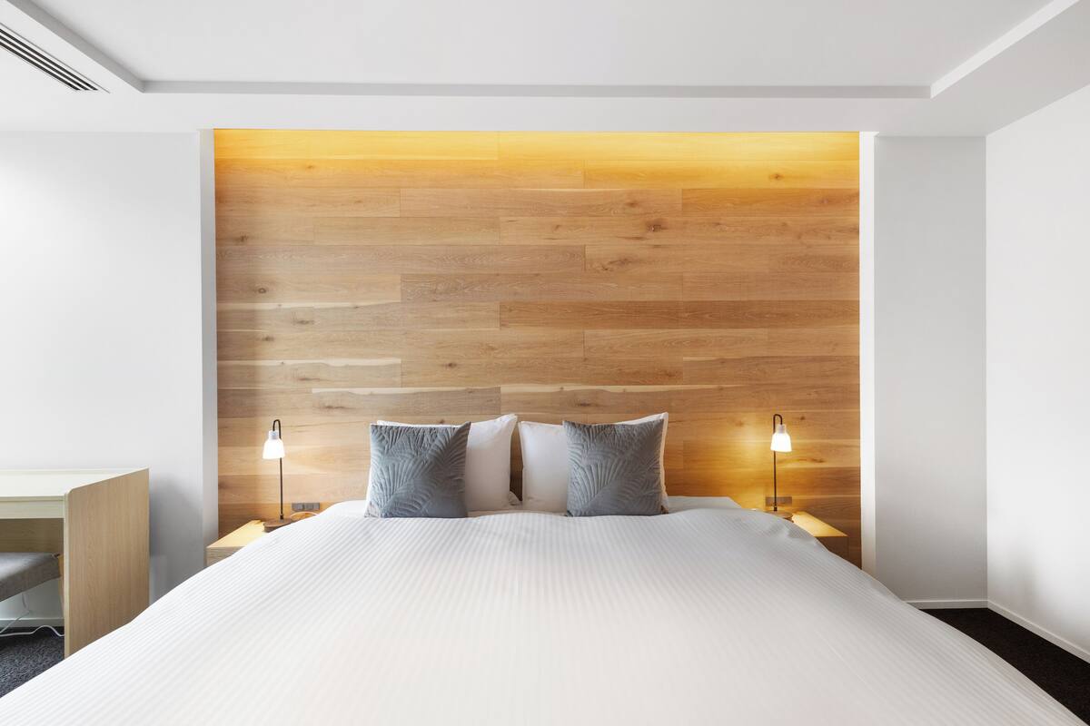 A modern hotel room with a wooden headboard, a bed with white linens, two bedside lamps, and a small desk with a chair.
