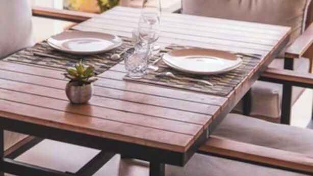 Mesa de madera preparada para dos personas, con una copa y un plato, una planta en maceta y vistas a vegetación exuberante.