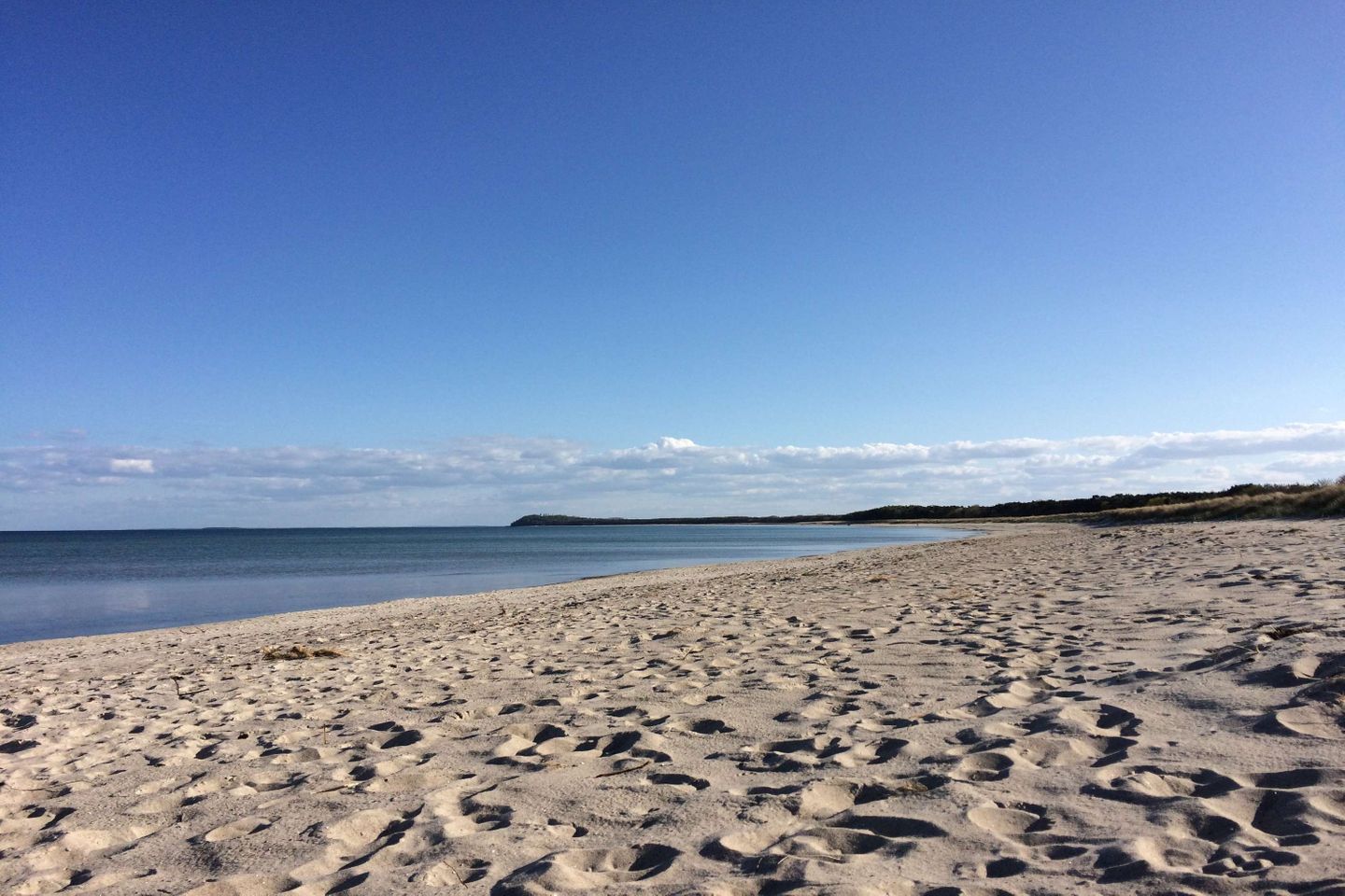 Une plage de sable où l’on peut voir des empreintes menant à l’eau, un ciel dégagé et une étendue de terre visible au loin.