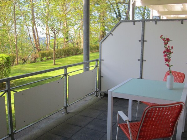 A balcony with a table and chairs, a vase with flowers, and a view of a green lawn and trees.