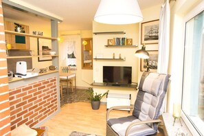 A cozy living room with a brick fireplace, a comfortable armchair, a television, and a bookshelf.