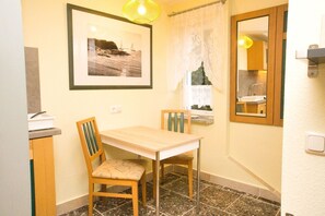 Ein kleiner Essbereich mit Holztisch und Stühlen, einem gerahmten Foto an der Wand, einem Fenster mit Spitzgittervorhängen und einem Spiegel über dem Tisch.