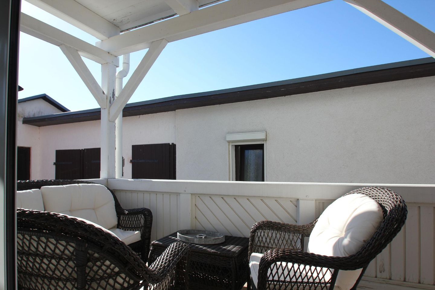 A covered outdoor seating area with wicker chairs and a small table, overlooking a building with a white exterior and dark shutters.
