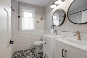 Ein Badezimmer mit weiß geflieste Wänden, einer weißen Toilette, einem Waschtisch mit Spüle und Spiegel sowie einem gemusterten Boden.