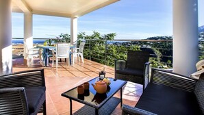 Uma área de estar coberta ao ar livre, com móveis de vime, mesa de vidro e vista para a costa.
