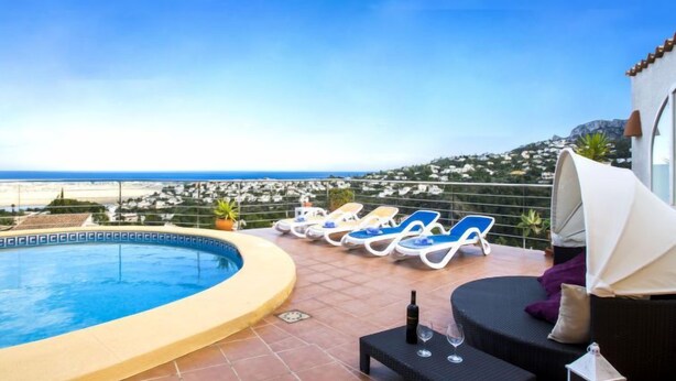 Uma área de piscina no terraço com vista para o mar e colinas, espreguiçadeiras, uma mesa pequena com garrafa e copos, e um espaço de estar coberto.