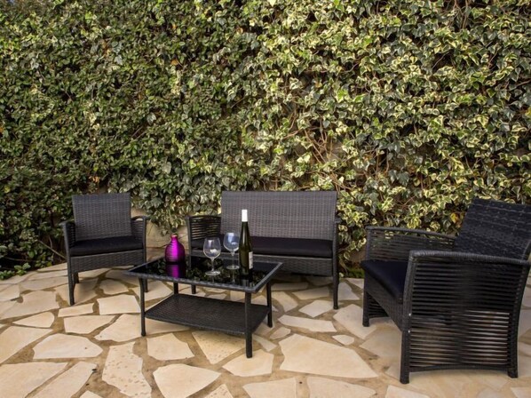 Un espace patio aménagé avec des meubles en osier et une table en verre, entouré d’un sol en pierre et d’un mur recouvert d’une épaisse végétation de lierre.