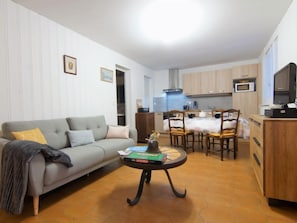 A compact living space with a sofa, a coffee table, a dining table with chairs, a kitchen area with cabinets and a microwave, and a television on a wooden cabinet.