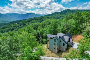 A two-story house with a stone foundation and a metal roof, surrounded by dense forest and mountains.