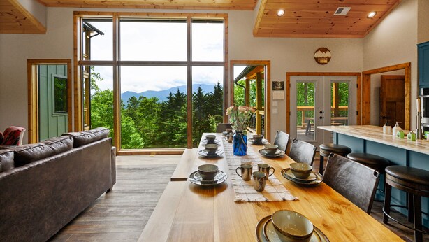 A spacious living room with a large wooden dining table, leather sofa, and a view of mountains through large windows. - Firefly Haus - Hot Tub, Firepit, 10min to Smokies (Gatlinburg)