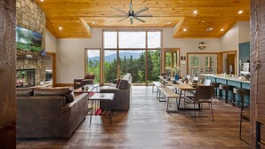 A spacious living area with a fireplace, a large window offering a mountain view, and a kitchen area with bar stools. - Firefly Haus - Hot Tub, Firepit, 10min to Smokies (Gatlinburg)